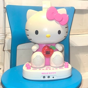 May include: A white Hello Kitty character with a pink bow and a red apple on her dress. The character is a speaker with two white speakers on the bottom and a control panel with the text "HELLO KITTY".