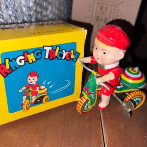 May include: A red and green wind-up toy tricycle with a smiling boy figure riding it. The toy is on a wooden surface and has a yellow box with the words "Ringing Tricycle" on it.