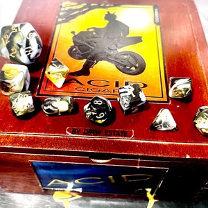May include: A wooden box with a black and white image of a person riding a motorcycle. The box is open and contains a set of black, white, and gold dice. The box has the words "Acid Cigar" and "By Drew Estate" printed on it.