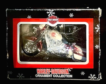 Puede incluir: Un adorno de motocicleta Harley-Davidson dentro de una caja rectangular negra con borde rojo y patrones de copos de nieve blancos. La caja presenta el texto Harley-Davidson Ornament Collection y el logotipo de Harley-Davidson arriba.