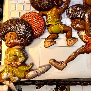 May include: Five vintage figurines of basketball players with afro hairstyles. The figurines are made of plastic and painted in brown, orange, yellow, and white. They are posed in various basketball-related actions.