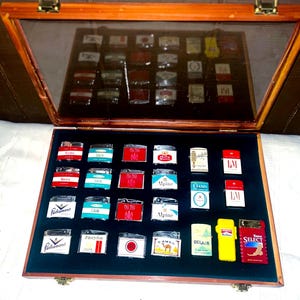 May include: A wooden display case with a glass lid, displaying a collection of vintage cigarette lighters. The lighters feature various brand logos and colourful designs, arranged on a dark blue velvet interior. The case is open, revealing the collection.