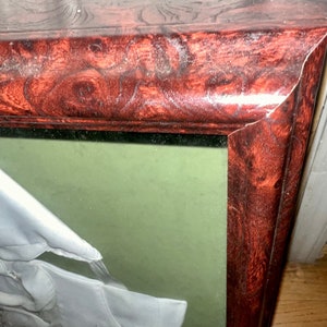 May include: A framed display of a white baby outfit with a green background. The frame is made of dark wood with a red-brown stain.