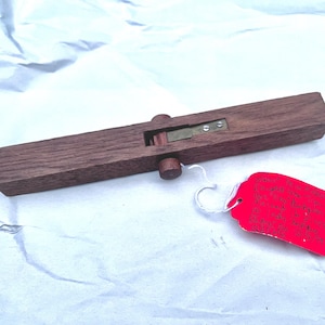 May include: A rectangular wooden tool with a brass blade and two wooden knobs. The tool is a dark brown color and has a red tag attached with white string. The tool is lying on a white surface.