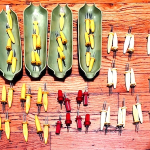 May include: Four elongated, light green corn-shaped serving dishes arranged vertically, accompanied by a collection of corn-on-the-cob holders with yellow, white, and red handles, each featuring two metal prongs for securing corn cobs.