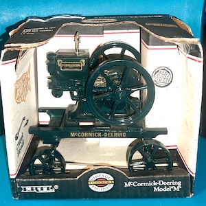 May include: A dark green McCormick-Deering Model M vintage gasoline engine model, displayed in its original box. The model features a flywheel, wheels, and the text "McCormick-Deering" and "Vintage Gasoline Engines". The box is white and black.