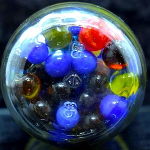 Può includere: Un barattolo di vetro trasparente pieno di biglie colorate. Le biglie sono di vari colori, tra cui blu, rosso, giallo e nero. Alcune biglie hanno i numeri "23" o "83" stampati. Il barattolo è su una superficie scura.