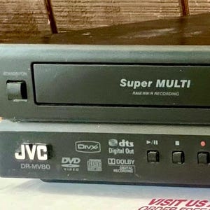 May include: A black JVC DR-MV80 DVD and VCR recorder featuring a disc tray labelled Super MULTI RAM/RW/R RECORDING. The front panel includes a power button, open/close button, and playback controls for play, stop, record, rewind, fast forward, and dubbing.