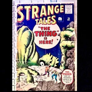 Puede incluir: Portada de cómic vintage de "Strange Tales" con temática de monstruos. La portada tiene un fondo azul con el título en amarillo y rojo. El texto "The Thing is Here!" se muestra con un monstruo verde y dos personas.
