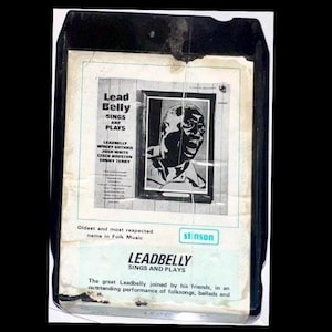 May include: A vintage black 8-track tape featuring the album "Leadbelly Sings and Plays." The album cover has a black and white portrait of Leadbelly. The text on the cover includes the album title and the names of the musicians.