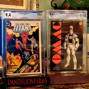 May include: Two graded comic books in protective cases. The New Teen Titans #1 graded 9.4 and Omac: One Man Army Corps #1 graded 9.4. Both comic books are displayed on a wooden shelf with a brown surface.