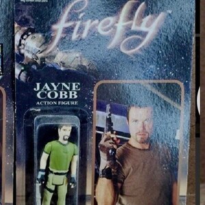 May include: A plastic action figure of Jayne Cobb from the TV show Firefly. The figure is wearing a green shirt, brown pants, and black gloves. The figure is packaged in a plastic blister pack with a cardboard backing. The backing features a photo of the actor who plays Jayne Cobb, Nathan Fillion, and the text "Firefly" and "ReAction Figures".