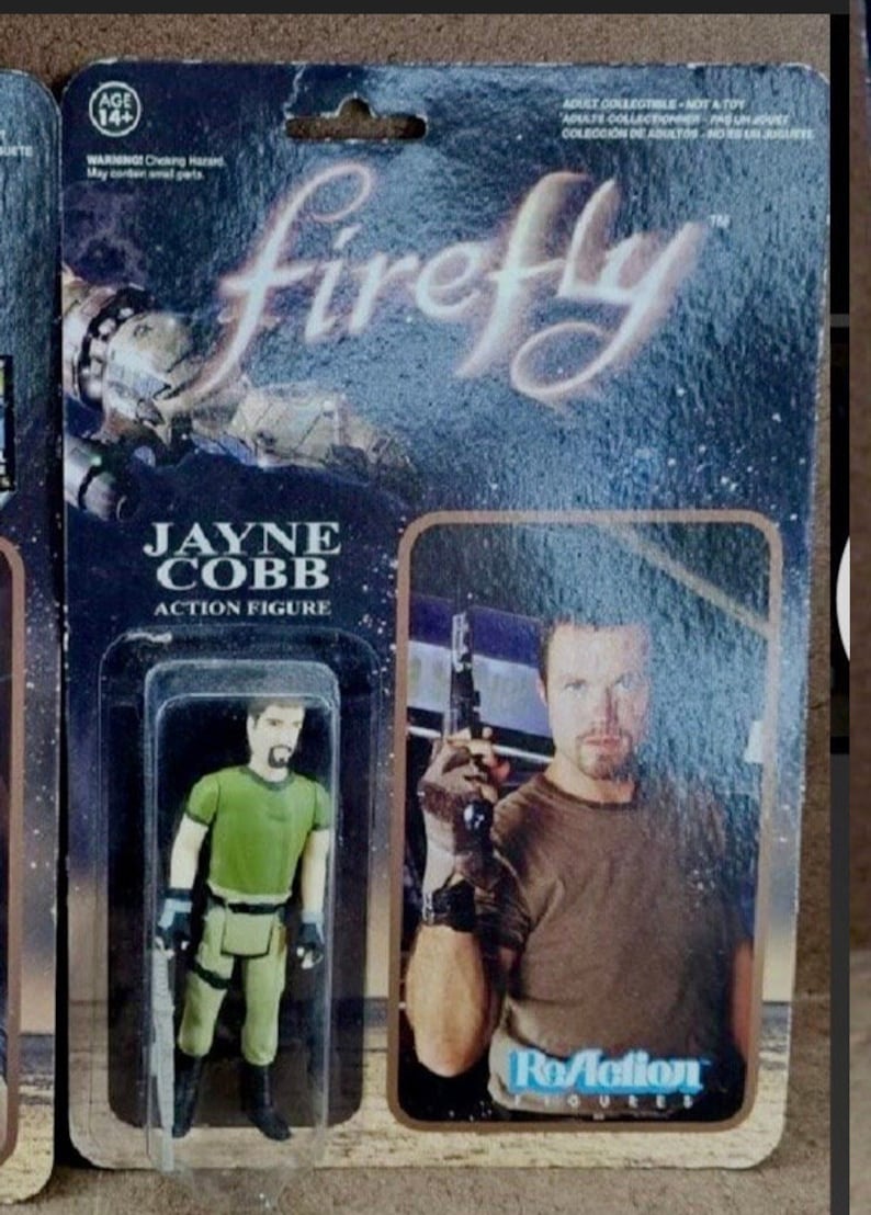 May include: A plastic action figure of Jayne Cobb from the TV show Firefly. The figure is wearing a green shirt, brown pants, and black gloves. The figure is packaged in a plastic blister pack with a cardboard backing. The backing features a photo of the actor who plays Jayne Cobb, Nathan Fillion, and the text "Firefly" and "ReAction Figures".