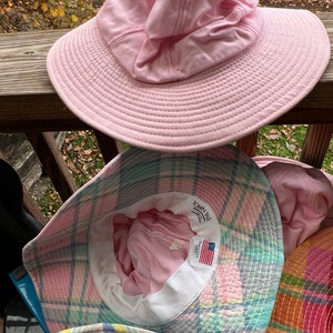 May include: A selection of sun hats in different colours and patterns. The top hat is a solid pale pink with a wide brim and a gathered crown. Below are hats with plaid and other colourful designs. The hats are made of fabric and are suitable for sun protection.