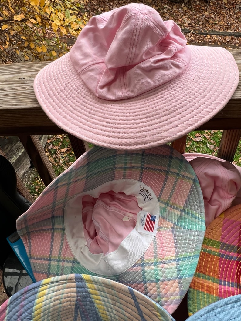 May include: A selection of sun hats in different colours and patterns. The top hat is a solid pale pink with a wide brim and a gathered crown. Below are hats with plaid and other colourful designs. The hats are made of fabric and are suitable for sun protection.