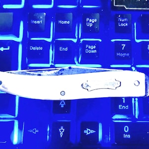 以下が含まれることがあります： 白柄と銀色の刃のポケットナイフが、青く照らされたキーボードの上に置かれています。ナイフには装飾的なエンブレムとリベットがあります。キーボードのキーには「Enter」や数字などのラベルが見えます。