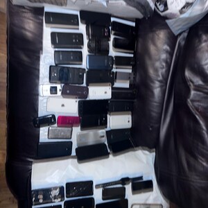 May include: A collection of various smartphones and mobile devices, in a variety of colors including black, white, and silver. The devices are arranged on a white surface, with a dark leather background.