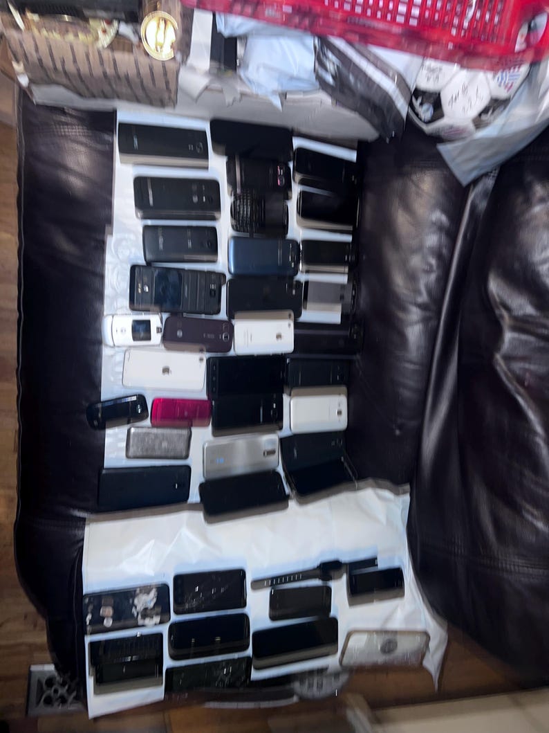 May include: A collection of various smartphones and mobile devices, in a variety of colors including black, white, and silver. The devices are arranged on a white surface, with a dark leather background.