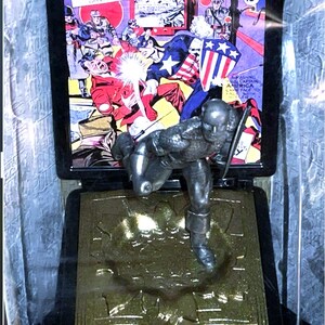 K&ouml;nnte beinhalten: Eine silberne Captain America-Figur in einer Vitrine. Dahinter ein Comic-Cover mit dem Titel "AMERICA COMICS" und dem Datum "M&Auml;RZ Nr. 1". Auf dem Sockel steht "CAPTAIN AMERICA 1941".