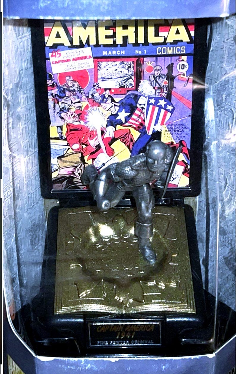 K&ouml;nnte beinhalten: Eine silberne Captain America-Figur in einer Vitrine. Dahinter ein Comic-Cover mit dem Titel "AMERICA COMICS" und dem Datum "M&Auml;RZ Nr. 1". Auf dem Sockel steht "CAPTAIN AMERICA 1941".