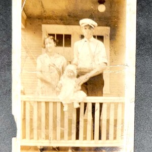 May include: Sepia-toned vintage photograph of a family posing on a porch. A woman and a man stand behind a baby seated on a railing. The photo is framed with a white border and shows signs of age.