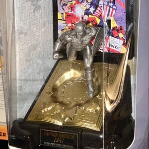K&ouml;nnte beinhalten: Captain America Figur aus feinem Zinn in einer Vitrine mit Comic-Cover-Hintergrund. Die Verpackung tr&auml;gt die Aufschrift "Comic Book Champions" und "Marvel Comics".