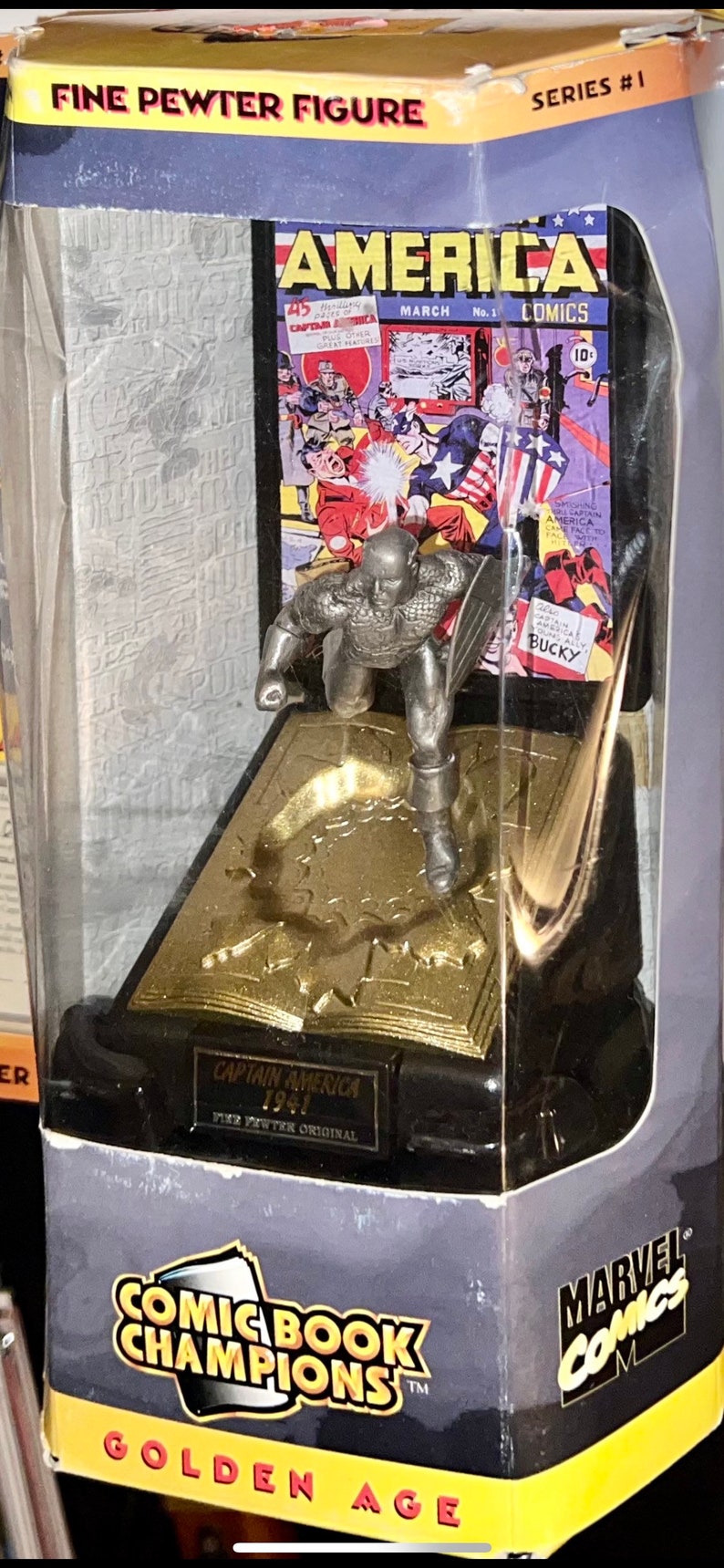 K&ouml;nnte beinhalten: Captain America Figur aus feinem Zinn in einer Vitrine mit Comic-Cover-Hintergrund. Die Verpackung tr&auml;gt die Aufschrift "Comic Book Champions" und "Marvel Comics".