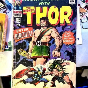 Op de afbeelding: Vintage stripboekcover voor "Journey Into Mystery" met "The Mighty Thor" en "Enter Hercules!". De illustratie toont een gespierde man met een wapen, Thor en een ander personage in gevecht. Het stripboek is gemarkeerd met "Marvel Comics Group" en "124 Jan 12".