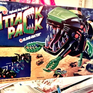 Puede incluir: Un camión de juguete verde y negro con una boca de monstruo y una función de disparo de slime. La caja dice "Attack Pack Slime-inator" y muestra el camión transformándose en un monstruo.
