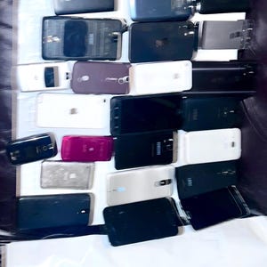May include: A collection of various smartphones and a smartwatch, in different colors and conditions. The phones are mostly black, white, and gray, with some showing signs of wear and tear. The image is taken from above, showing the devices arranged on a white surface.