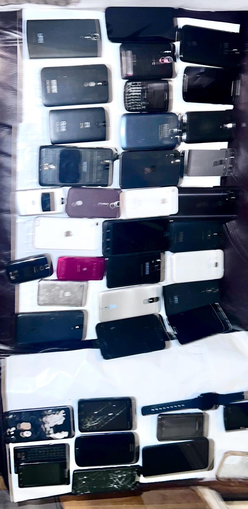May include: A collection of various smartphones and a smartwatch, in different colors and conditions. The phones are mostly black, white, and gray, with some showing signs of wear and tear. The image is taken from above, showing the devices arranged on a white surface.