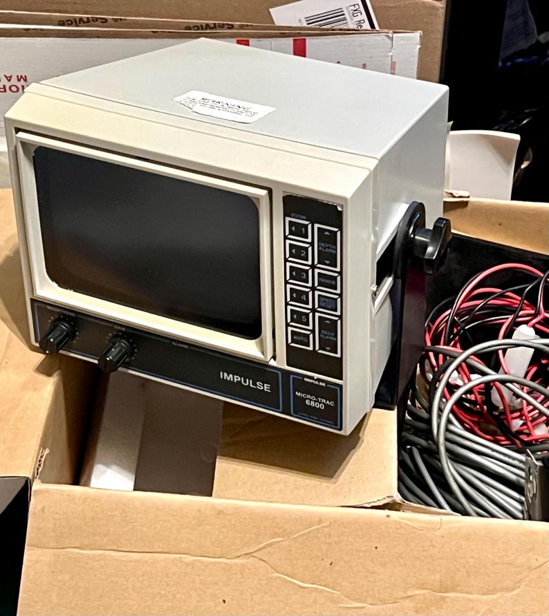 May include: An off-white Impulse Micro-Trac 6800 marine electronic display unit with a dark screen, black control knobs, and a vertical keypad. The device features a black mounting bracket and is accompanied by a bundle of red, black, and gray wires.