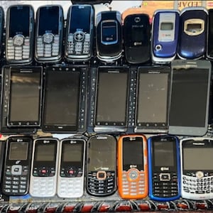 May include: A collection of various cell phones, including flip phones and smartphones, arranged in multiple rows. The phones display a range of colors, including black, silver, blue, and orange. Some phones have visible keypads, while others have touchscreens.
