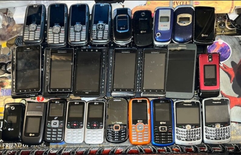 May include: A collection of various cell phones, including flip phones and smartphones, arranged in multiple rows. The phones display a range of colors, including black, silver, blue, and orange. Some phones have visible keypads, while others have touchscreens.