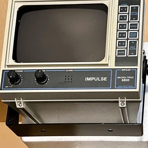 May include: A gray Impulse Micro-Trac 6800 video fish finder with a dark screen, two black control knobs, and a side panel featuring buttons labeled zoom, depth alarm, range, speed reset, and deep alarm, all mounted on a black metal bracket.
