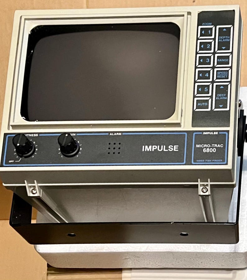 May include: A gray Impulse Micro-Trac 6800 video fish finder with a dark screen, two black control knobs, and a side panel featuring buttons labeled zoom, depth alarm, range, speed reset, and deep alarm, all mounted on a black metal bracket.