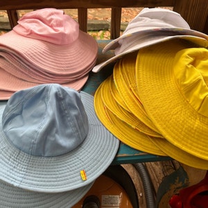May include: A selection of sun hats in shades of pink, yellow, white, and light blue. The hats feature wide brims and a traditional design, ideal for shielding from the sun. The hats are arranged on a wooden surface.