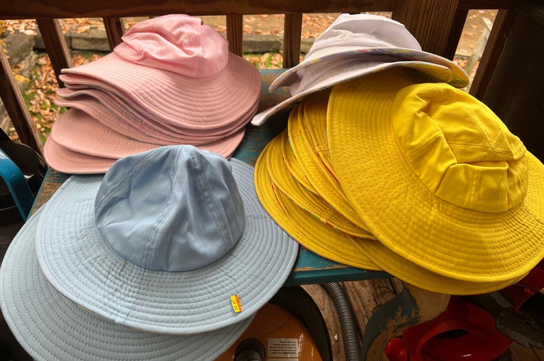 May include: A selection of sun hats in shades of pink, yellow, white, and light blue. The hats feature wide brims and a traditional design, ideal for shielding from the sun. The hats are arranged on a wooden surface.