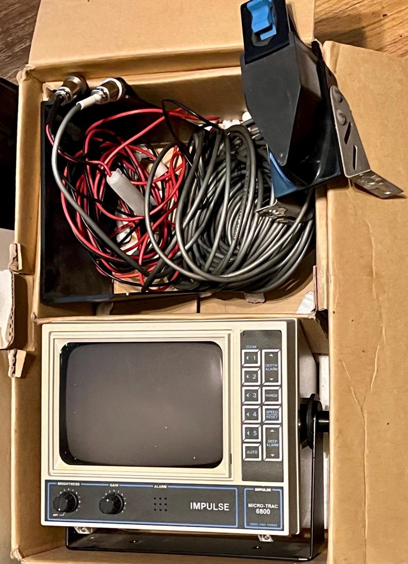 May include: An Impulse Micro-Trac 6800 fish finder unit with a beige casing, a dark screen, and a control panel featuring buttons and knobs. The unit is packed in a cardboard box with a bundle of gray and red electrical cables and a black transducer.