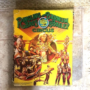 Peut inclure: Un programme souvenir de 1989 du cirque Ringling Bros. and Barnum & Bailey avec une couverture jaune vif. La couverture présente des illustrations d'un clown, d'un léopard, d'un tigre et d'acrobates, avec le texte The Greatest Show on Earth.