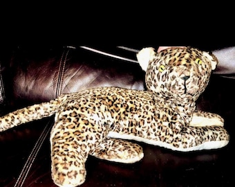 Puede incluir: Un peluche de leopardo con un patrón de pelaje moteado en color canela y negro, ojos amarillos y una nariz negra cosida. El animal de peluche se muestra en posición tumbada con la cola extendida, con un tejido exterior suave y texturizado.