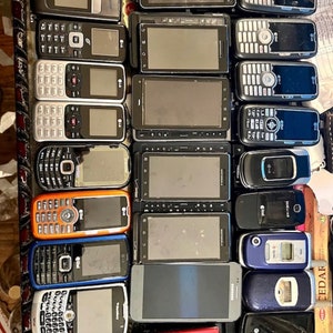 May include: A collection of various used cell phones, including flip phones and smartphones, in different colors and designs. The phones have visible screens, keypads, and brand logos. Some phones are black, orange, blue, and silver.
