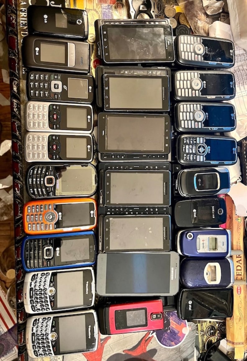 May include: A collection of various used cell phones, including flip phones and smartphones, in different colors and designs. The phones have visible screens, keypads, and brand logos. Some phones are black, orange, blue, and silver.