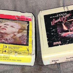 May include: Two vintage David Bowie 8-track tapes. One features the album "Huney Dory" with a portrait of Bowie. The other is "Young Americans" with a pink and black album cover. Both are in off-white cases with RCA logos.