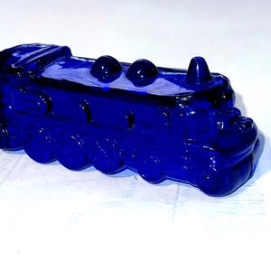 May include: A deep blue glass train ornament. The toy train features a detailed design with wheels, a chimney, and three circular details on top. The train is a solid, vibrant blue colour and is likely a decorative item.