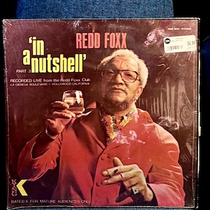 以下が含まれることがあります： レッド・フォックスのアルバム「In a Nutshell, Part 3」のジャケット。赤いジャケットを着たレッド・フォックスが、グラスとタバコを持っています。アルバムには「Redd Foxx Club, La Cienega Boulevard, Hollywood, California」と記載されています。