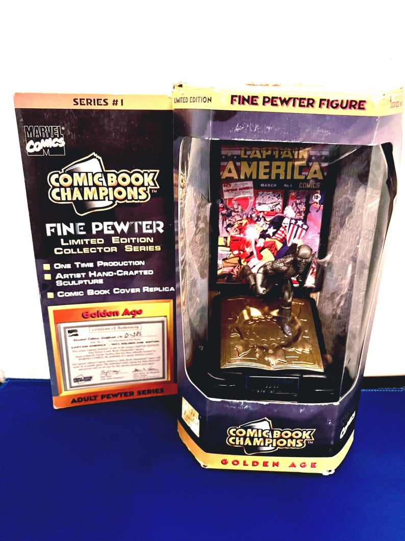 K&ouml;nnte beinhalten: Eine limitierte "Comic Book Champions" Feinzinnfigur von Captain America, pr&auml;sentiert in einer transparenten Kunststoffbox. Die Figur steht auf einem goldfarbenen Sockel mit offenem Buchdesign. Die Verpackung enth&auml;lt den Text "Golden Age" und "Marvel Comics".