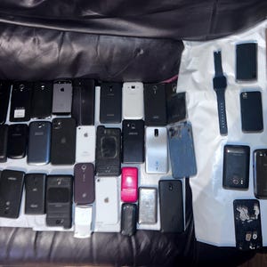 May include: A collection of various smartphones and a smartwatch, in different colors and designs, are arranged on a white surface. The phones vary in size and shape, with some featuring visible screens and others with their backs facing up.