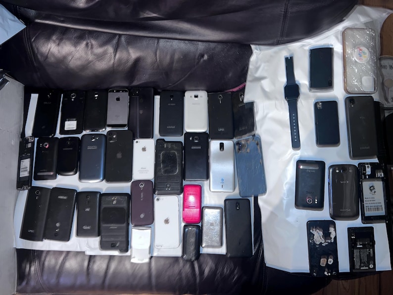 May include: A collection of various smartphones and a smartwatch, in different colors and designs, are arranged on a white surface. The phones vary in size and shape, with some featuring visible screens and others with their backs facing up.