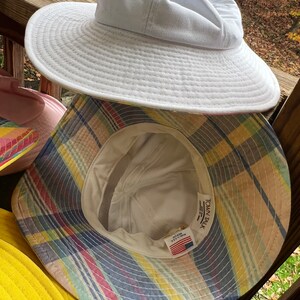 May include: A selection of sun hats in different colours. The top hat is white with a wide brim. Beneath it is a plaid bucket hat with yellow, blue, and pink stripes. Other hats in pink and yellow are also visible.